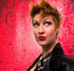 A white woman in a black leather jack with red lipstick and a pixie cut looks off to the side.